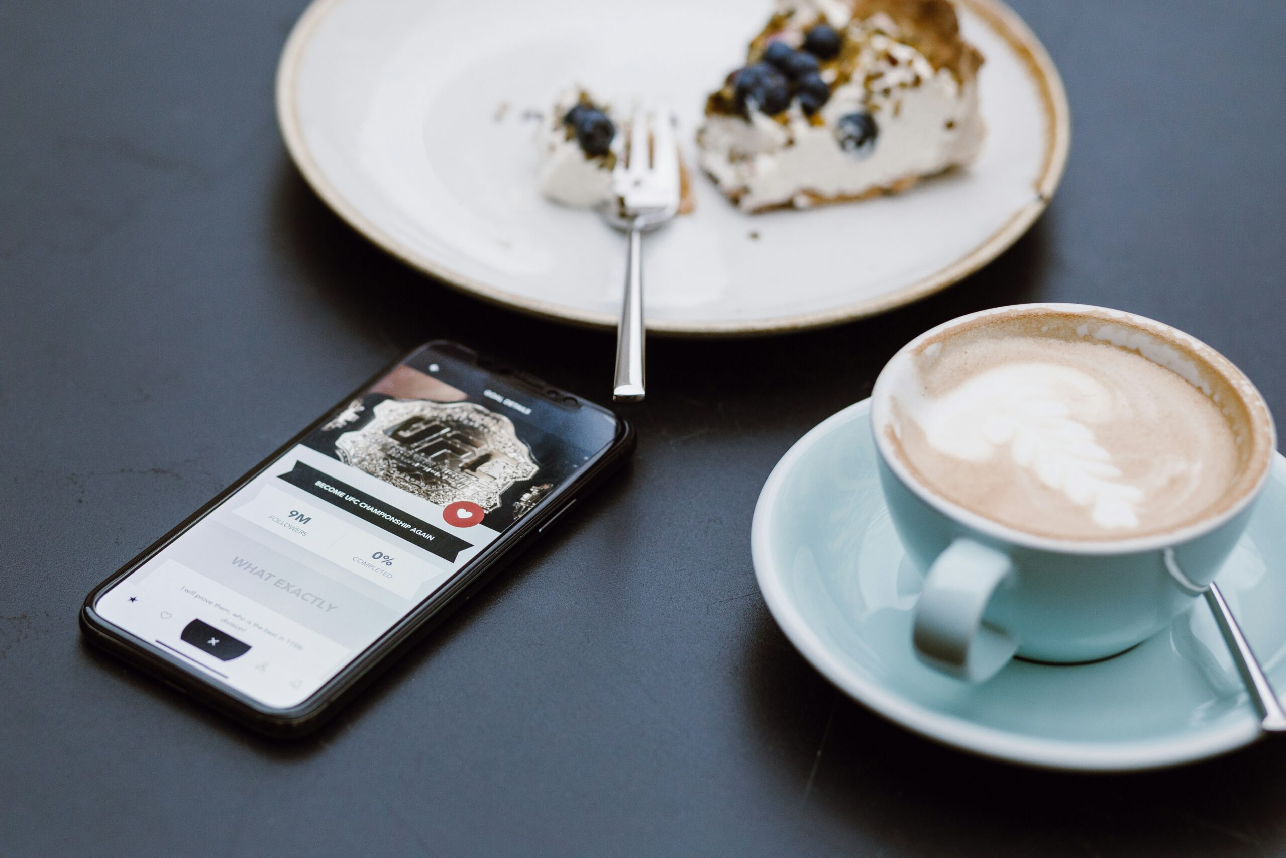 Teller mit einem angefangenem Stück Kuchen. Links daneben liegt ein Handy und rechts steht eine Kaffeetasse