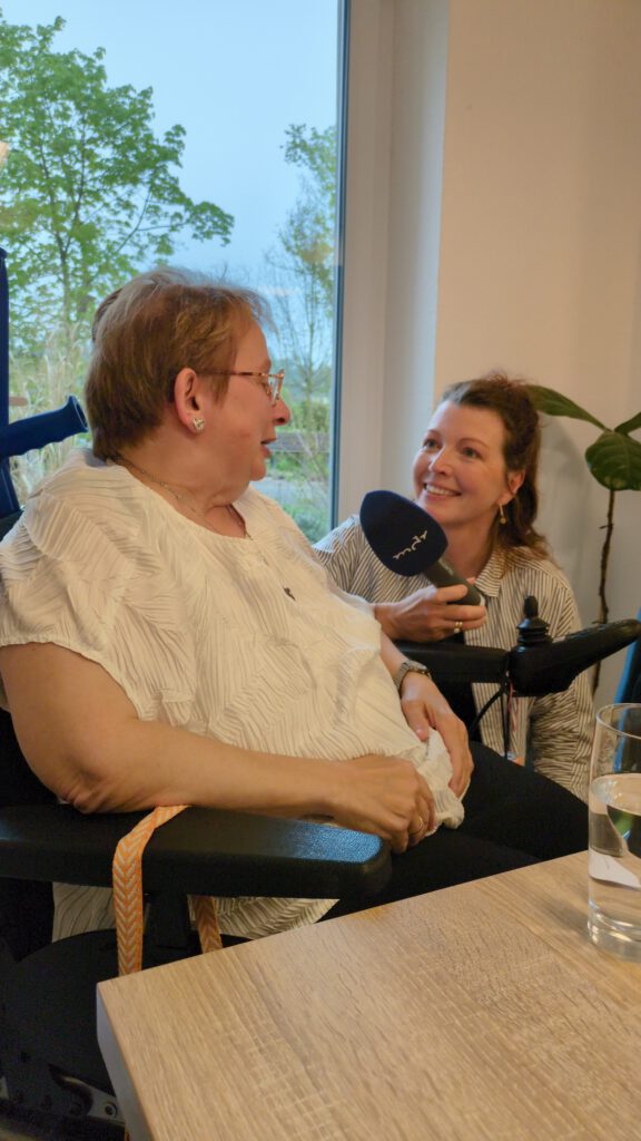 Eine Frau im Rollstuhl wird von einer jungen Frau mit Mikrofon in der Hand interviewt